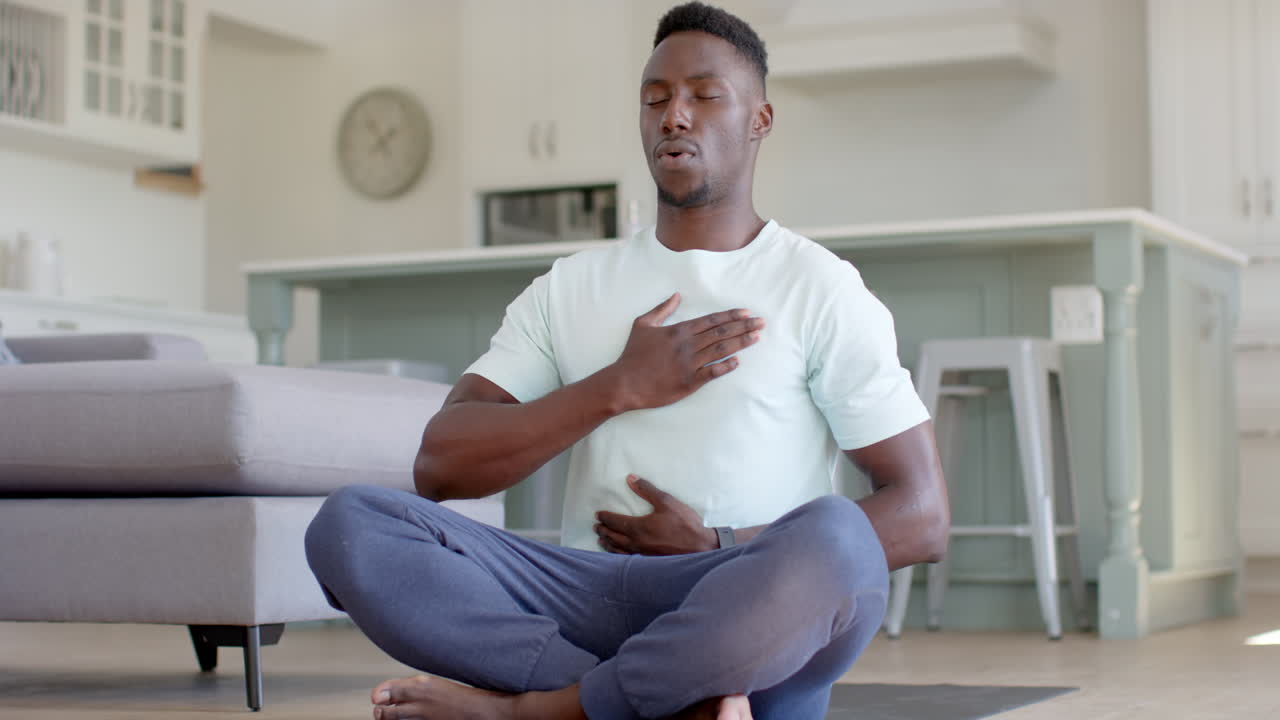 hombre afroamericano concentrado practicando meditación de yoga en la sala de estar soleada, cámara lenta