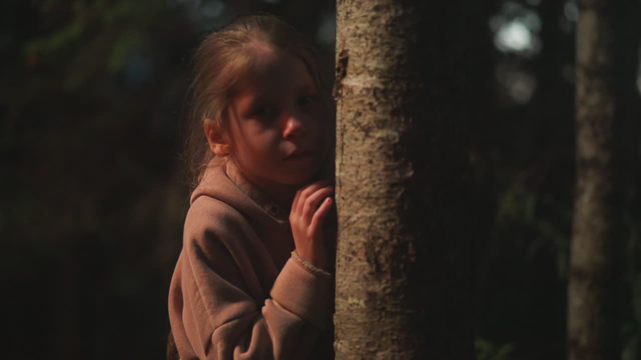 niño solitario se apoya en el tronco de un árbol en el bosque oscuro. niña pequeña perdida en el parque natural nocturno durante el viaje. niño amenazado por la oscuridad y los sonidos en el bosque salvaje