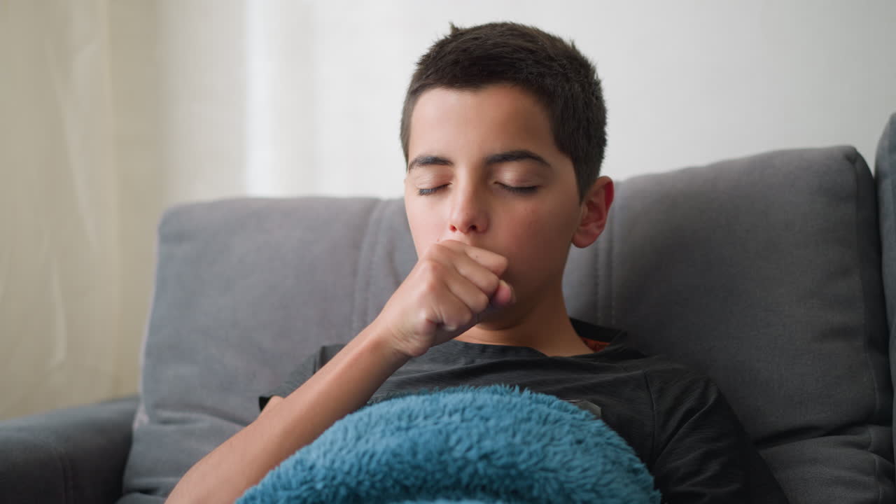 Portrait view of young boy lying on couch covered with blue towel coughing and showing signs of flu while adjusting position, with expression of discomfort, and illness in calm home setting