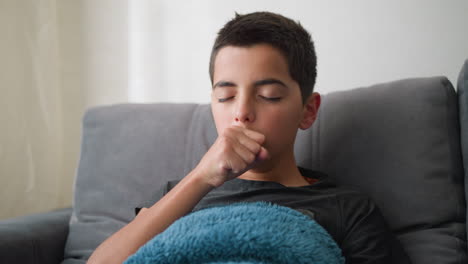 portrait view of young boy lying on couch covered with blue towel coughing and showing signs of flu while adjusting position, with expression of discomfort, and illness in calm home setting