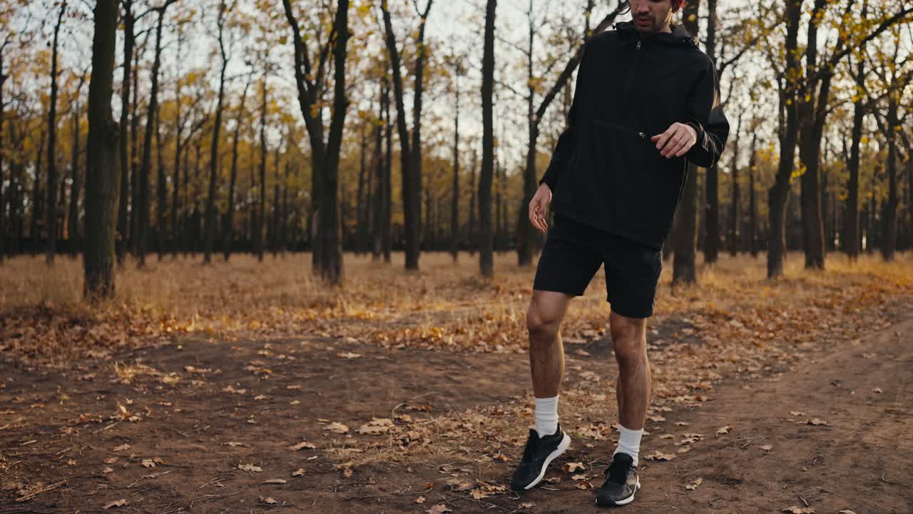 A brunette man in a black sports uniform and a red hat stretches his injured leg while jogging in the autumn forest in the morning along an earthen path