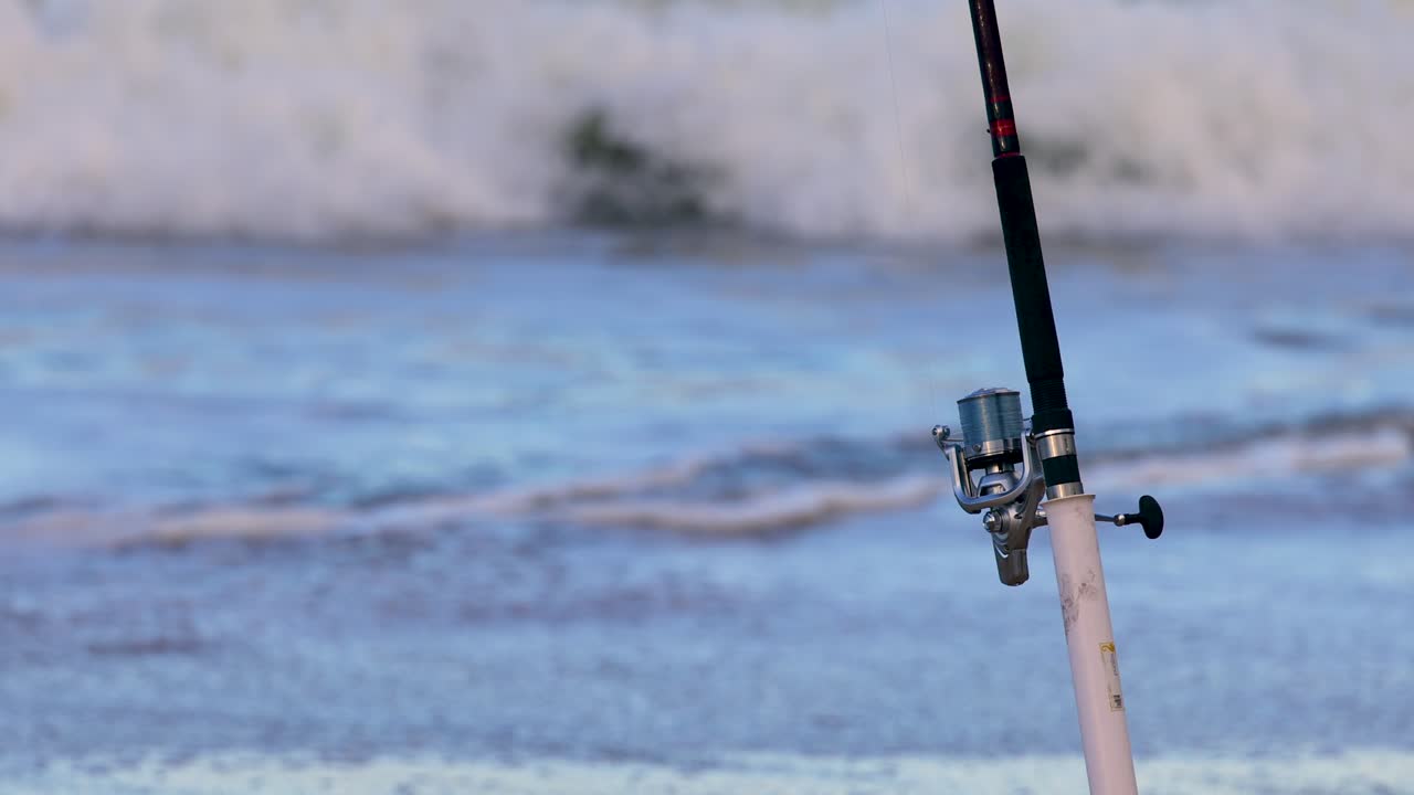 A fishing rod stands against rolling ocean waves at Charlesworth Bay Beach, Coffs Harbour, under natural daylight