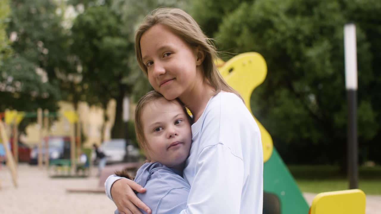 vista en primer plano de una niña con síndrome de down abrazando a otra niña mientras miran a la cámara en el parque en un día ventoso