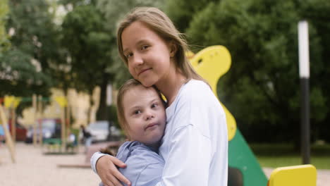 Close-up-view-of-a-little-girl-with-down-syndrome-hugging-another-girl-while-they-looking-at-camera-in-the-park-on-a-windy-day