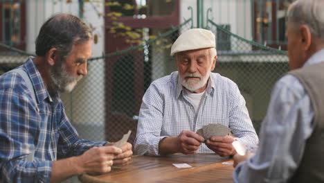 Three-Senior-Men-Best-Friends-On-Retirement-Playing-Cards-In-The-Yard-At-The-Table