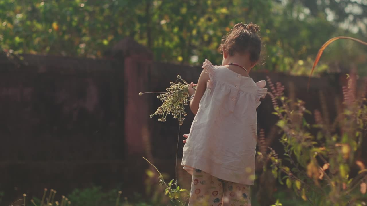 toma estática de mano de una joven india con flores en la mano parada en un jardín con plantas, árboles y una pared en el fondo mientras tira una planta