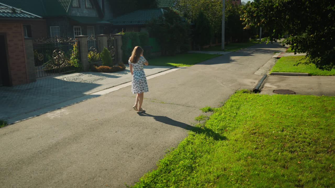 Lady in summer dress walks along quiet residential street, gently removing something from her eye as butterfly flutters nearby, under bright sunlight with shadow stretching across pavement