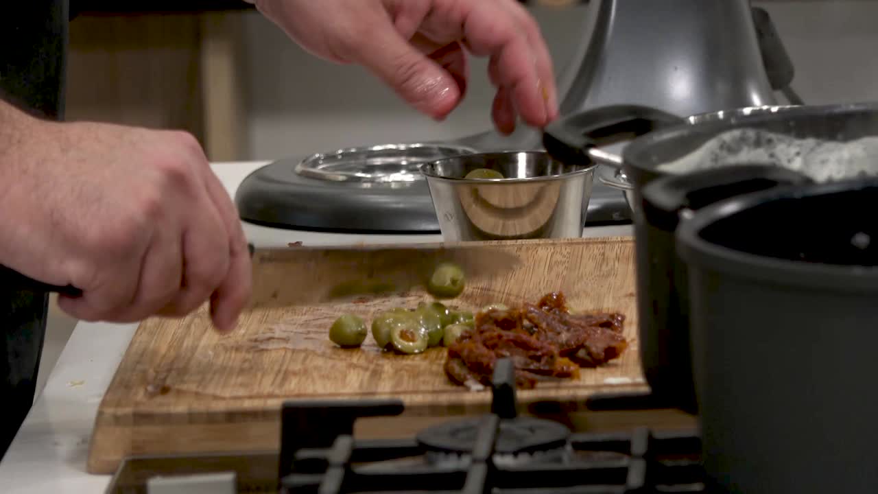 A chef skillfully slices green olives while adding spices and seasonings to a wooden cutting board. The vibrant kitchen is set for an exciting cooking experience