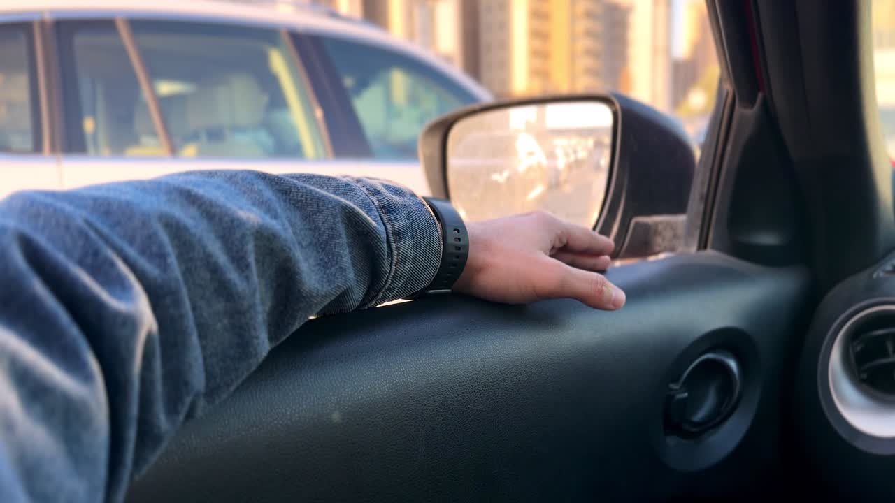 Nærbilde av en persons hånd som hviler på et bildørpanel inne i et kjøretøy, med sidespeil som reflekterer trafikk og bybygninger under varmt solnedgangslys under bykjøring.