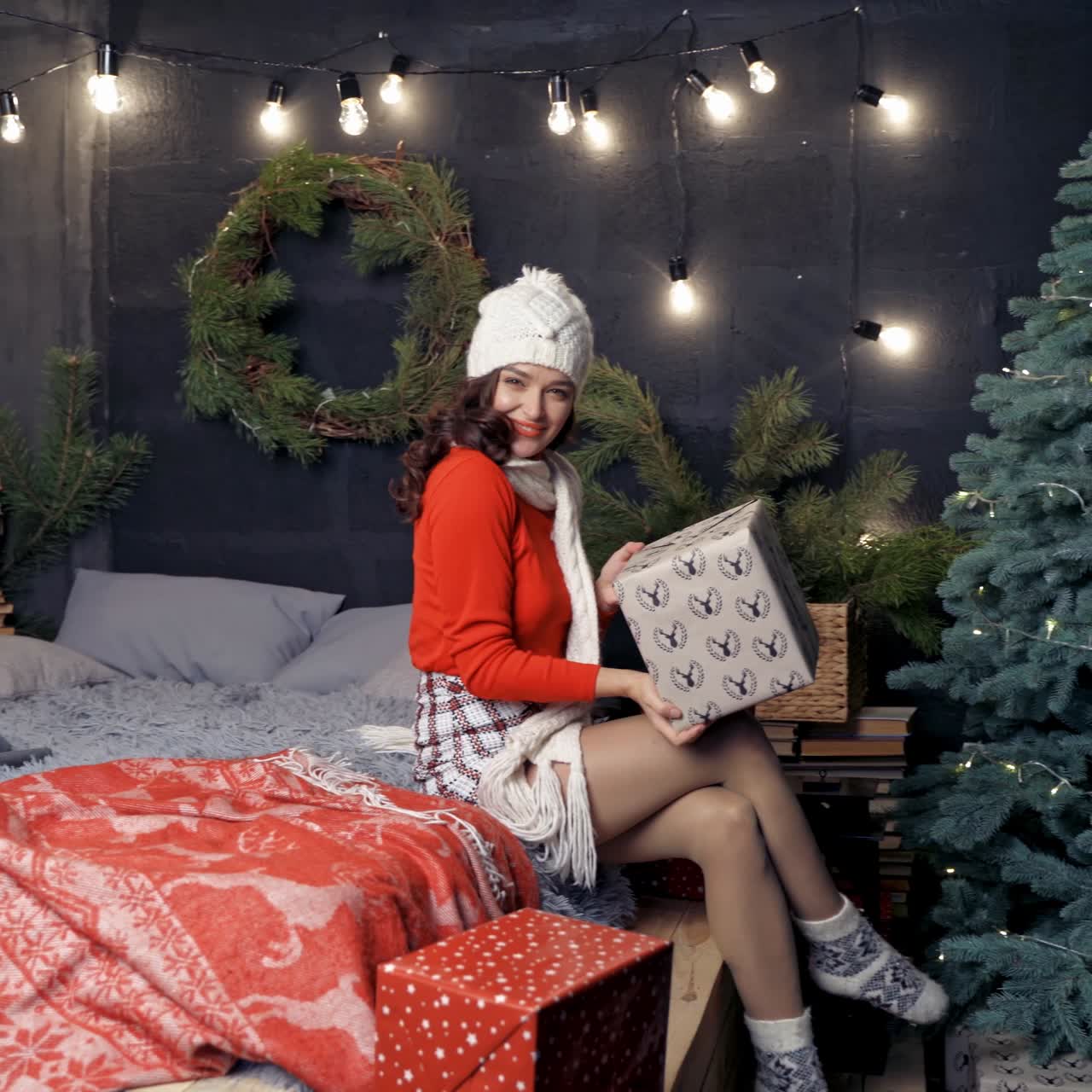 Positive young woman with gift box. Attractive girl in knitted cap and scarf sitting on bed near the Christmas tree with a big present and throwing it happily. Happy New Year.