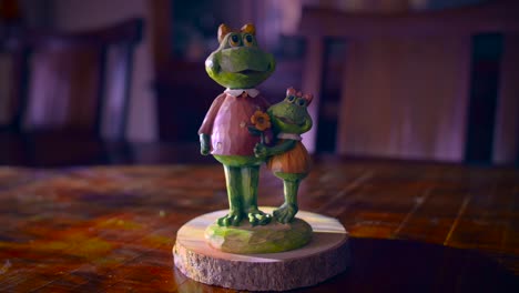 stone figurine of a mother and daughter frog standing on a stone base. the figurine is on a wooden tree trunk stand. the young frog is holding a flower and the mom is happy and smiling.