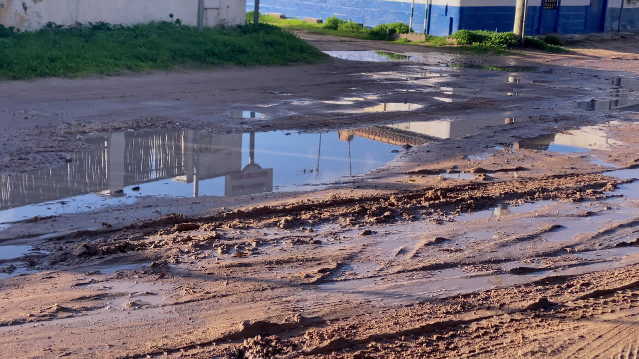 Orme di fango e pozzanghere di pioggia riflettono la delicata luce del mattino su una strada di accesso costiera sabbiosa dopo la pioggia. Una spiaggia sabbiosa si apre tra basse dune con tracce di pneumatici che conducono verso le onde dell'oceano
