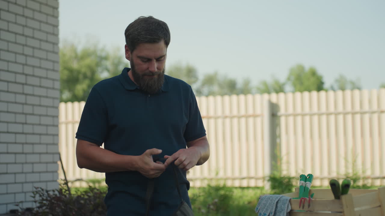 Young man gardener in yard picks apron belt, loops strap around waist while adjusting fit beside house fence, blue drum with crate, gloves, trowel, fork, sprayer nearby