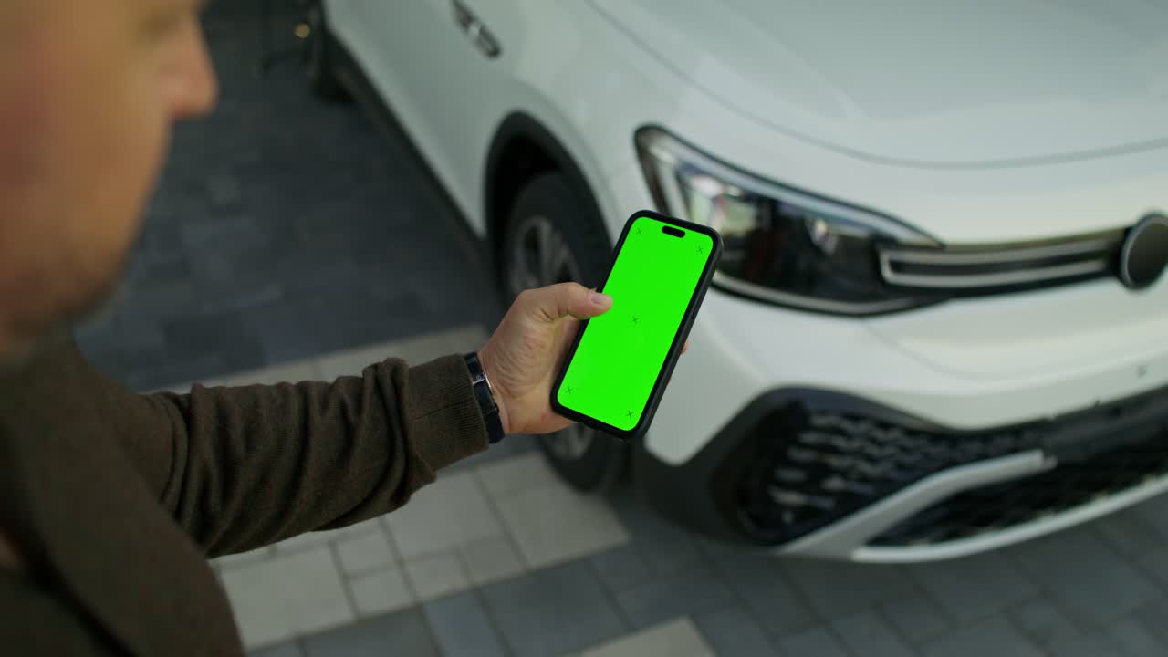 hombre sosteniendo un teléfono inteligente con pantalla verde frente a un coche blanco