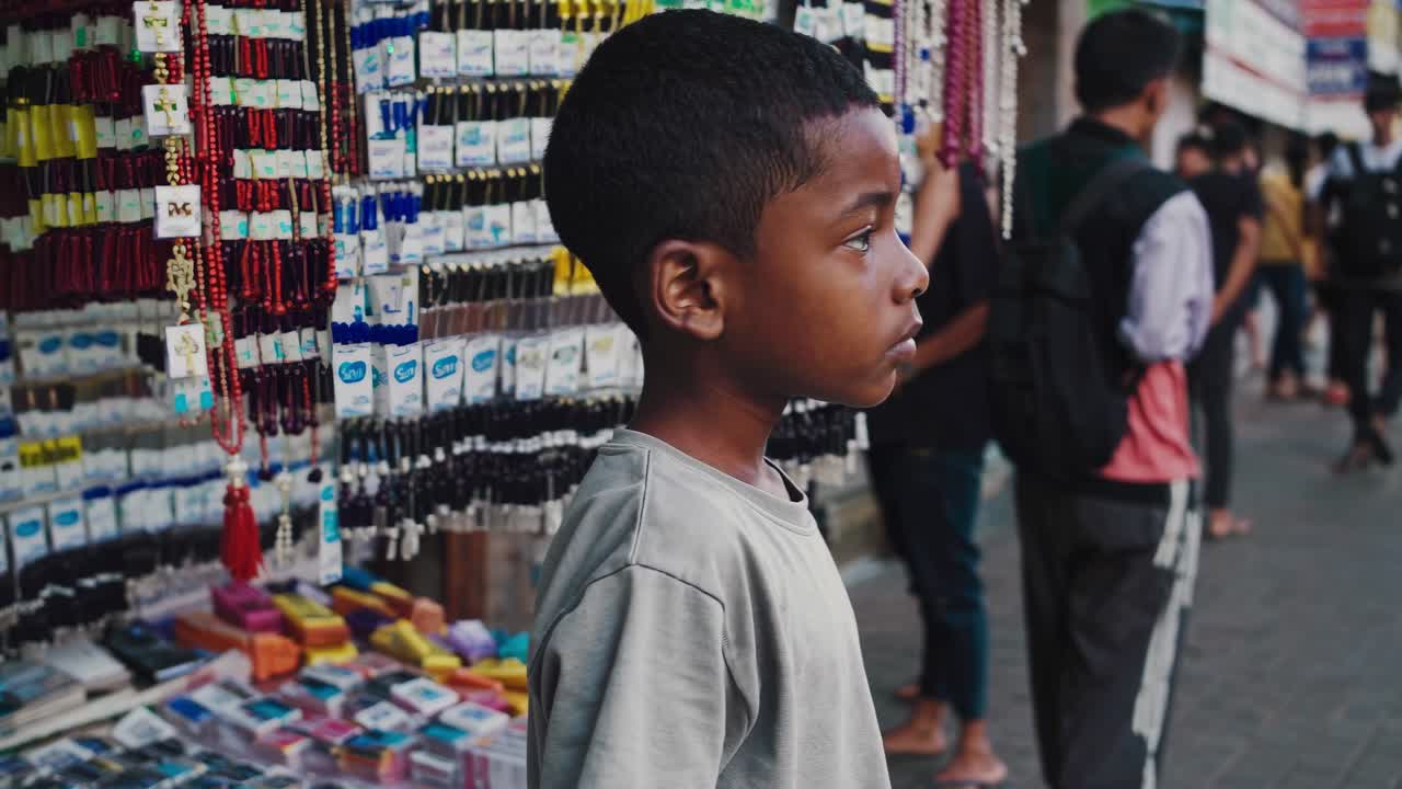 Young boy standing beside a colorful street stall filled with small items, observing the bustling activity of a busy urban street in an Asian city