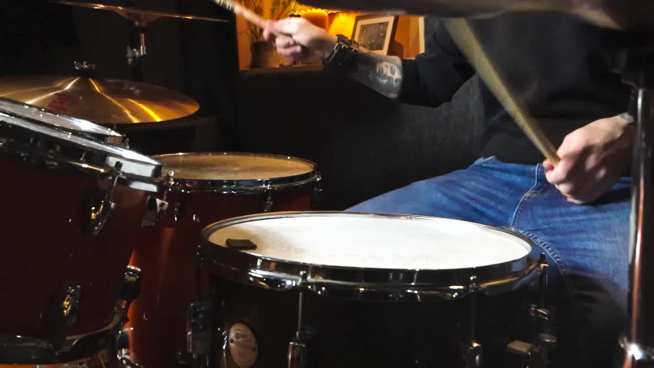 Gorgeous static slow motion footage of a man playing acoustic drums and hitting bronze and orange colored shiny drum cymbals next to him. The man has tattoos and is playing in a well lit studio room.