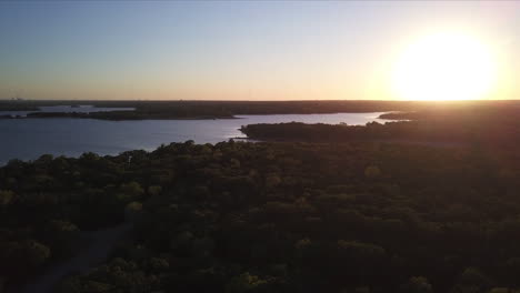 Backward-establishing-shot-of-a-dense-forest-beside-a-lake-at-sunset