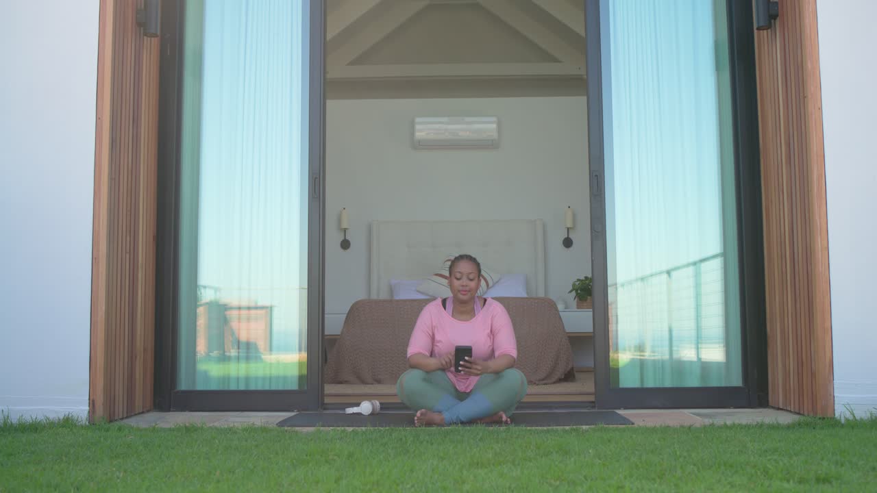 African American woman scooting on patio yoga mat after door opening donning headphones relaxing