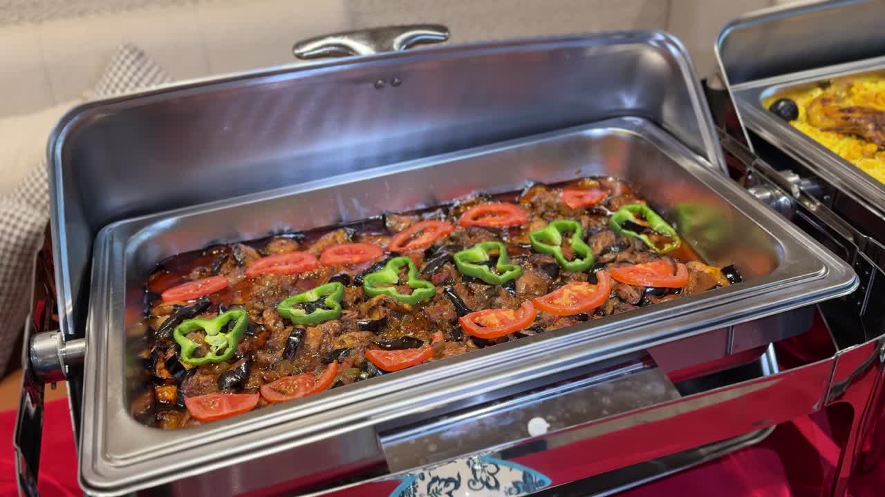 Primo piano di uno scaldavivande da buffet ripieno di verdure cotte, melanzane, pomodori e peperoni verdi in salsa saporita, esposto su un tavolo da catering al chiuso durante un evento di ristorazione.