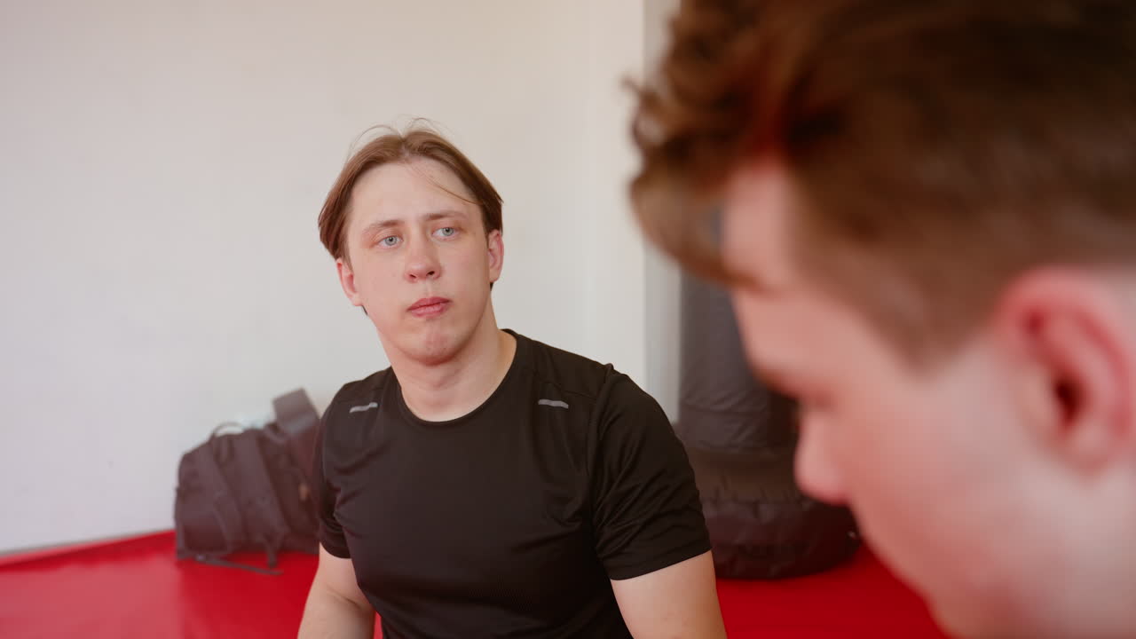 Athlete in black sportswear sits on red mat inside gym, looking with focused expression at training partner, showing intensity, determination, and concentration during boxing preparation