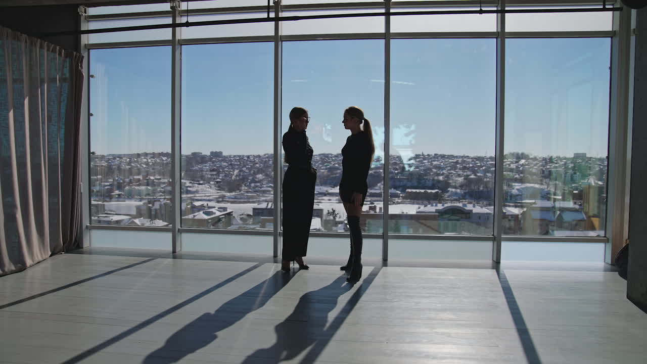 Friendly conversation between two female employees. Women in black stand near the windows, talk and then look at the cityscape.