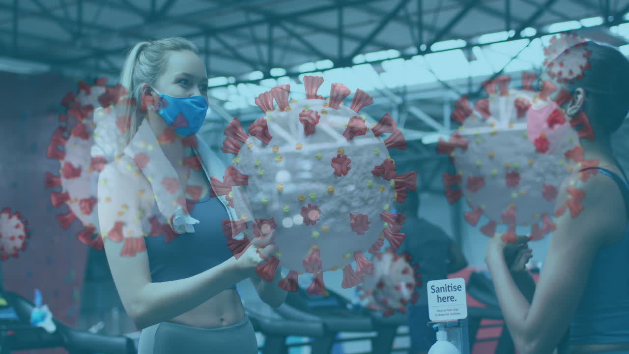 Covid-19 cells floating against two women wearing face masks sanitizing their hands at the gym