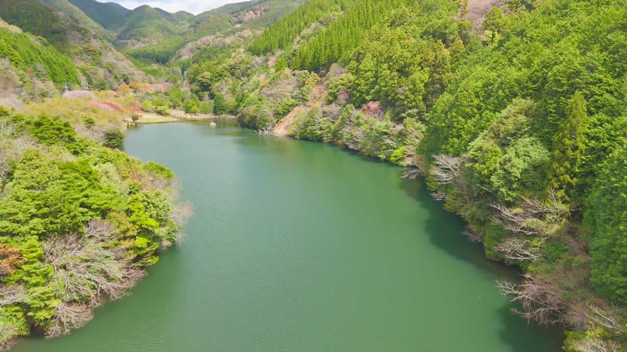 A stunning aerial shot flying over a small dam's spillway, gliding above the calm green reservoir to reveal the vast, lush mountain valley and forest ahead