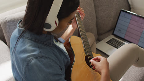 Video-of-happy-biracial-woman-in-headphones-sitting-on-sofa-and-playing-guitar