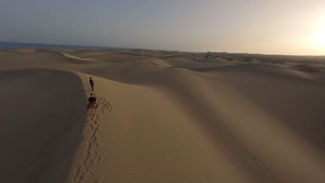 Antena-Dando-Vueltas-A-Personas-Irreconocibles-En-La-Cima-De-Una-Duna-De-Arena-Al-Atardecer,-Maspalomas