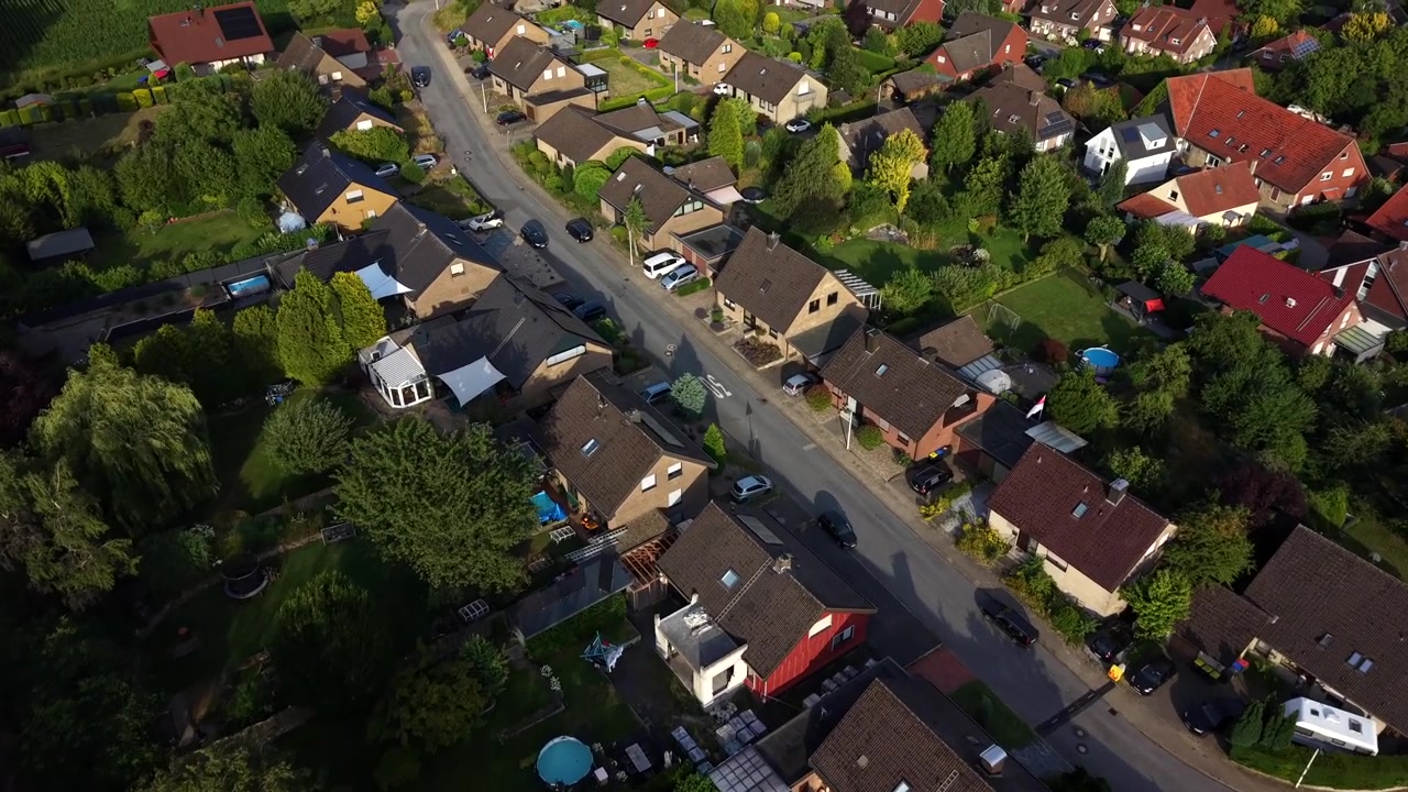 Gouden zonsonderganglicht baadt puntgevels in een rustige Duitse buitenwijk, met ronde opbouwzwembaden verspreid in tuinen langs een met bomen omzoomde woonstraat in een landelijke dorpsomgeving. Luchtfoto met drone
