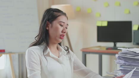 지친 아시아 여성이 사무실에서 문서와 함께 열심히 일하면서 머리를 흔들고 있습니다.