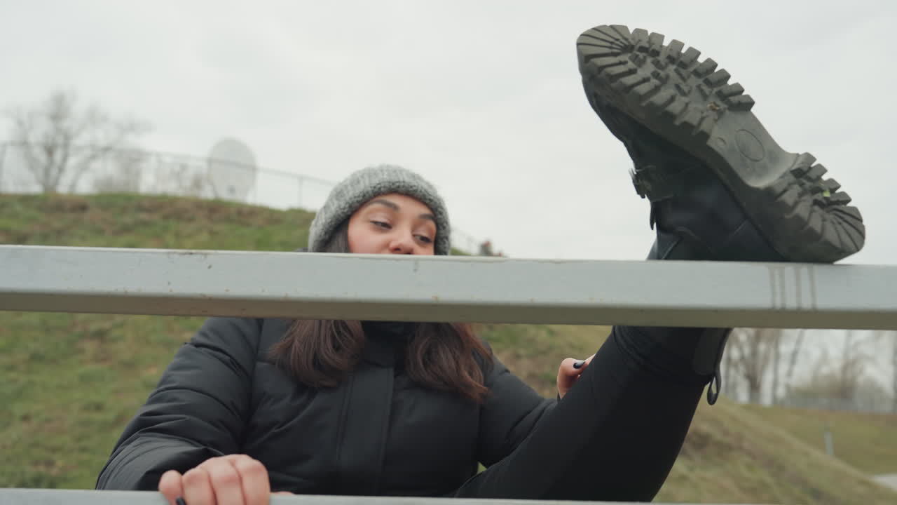 Mujer levantando la pierna sobre la barandilla, detalle de la suela de la bota y expresión concentrada, colina de parque al aire libre bajo cielo nublado, abrigo de invierno y gorro de punto, movimiento de baile improvisado que combina estiramiento y juego