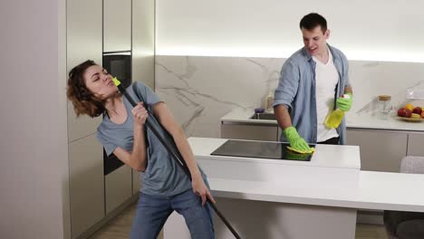 two young people dancing together and emotionally singing using broom and cleaning tools as as microphones while cleaning in kitchen. happy couple enjoying time, feel like rock stars