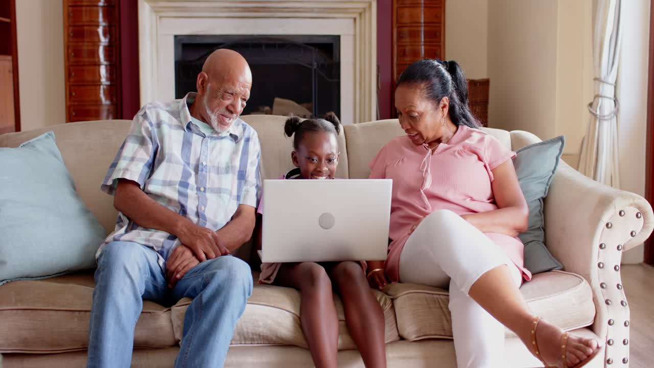 Diverse grandparents and granddaughter using laptop on sofa, sharing joyful moments, at home