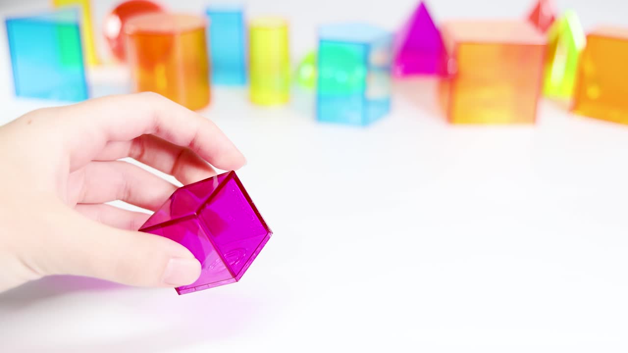 Hands manipulate vibrant 3D geometric shapes on a white table, showcasing various forms and colors in bright lighting