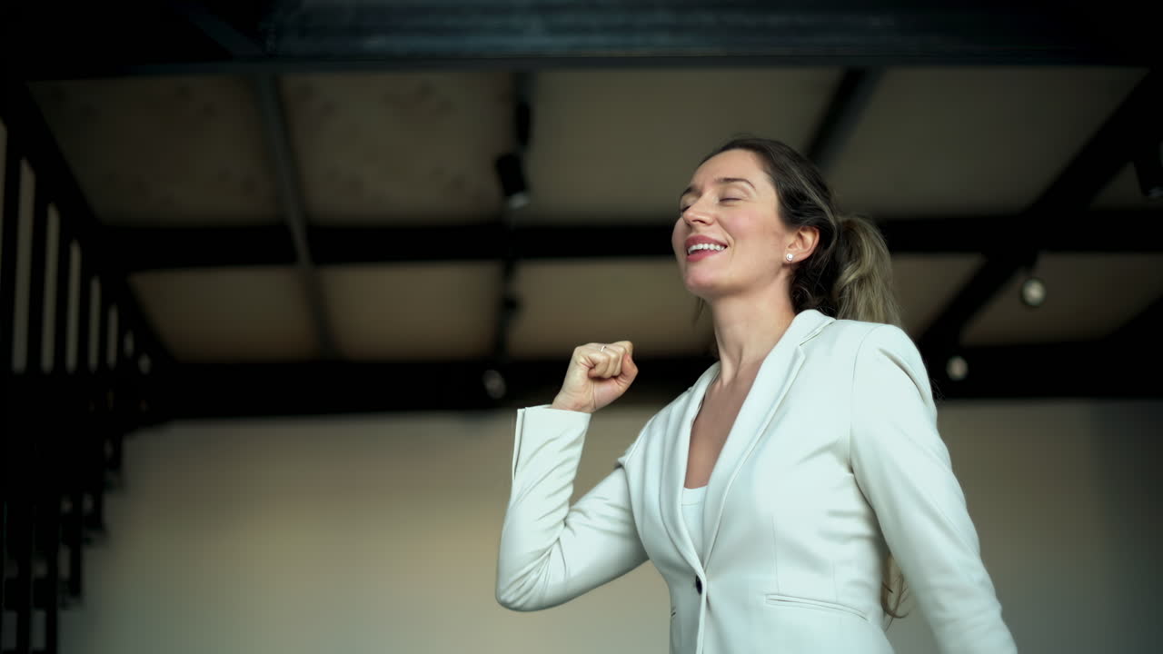 A joyful woman in a white blazer raises her arm in celebration inside a modern office. Her bright smile reflects happiness and achievement in a professional environment