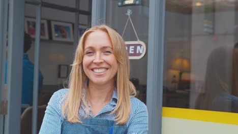 portrait of female owner or staff outside shop or cafe standing by open sign in doorway