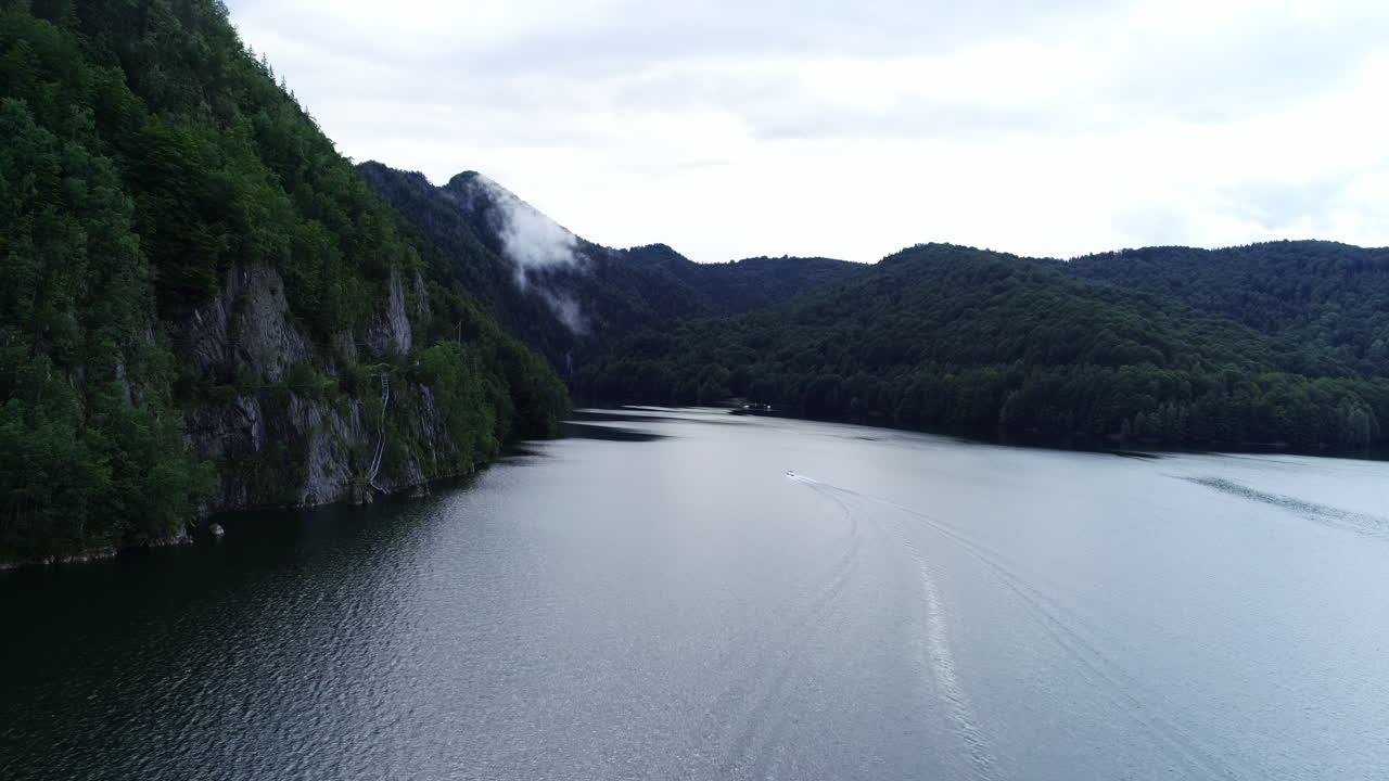 A tranquil lake reflects the early morning light, surrounded by lush mountains and gentle mist. A boat glides across the calm water, creating soft ripples