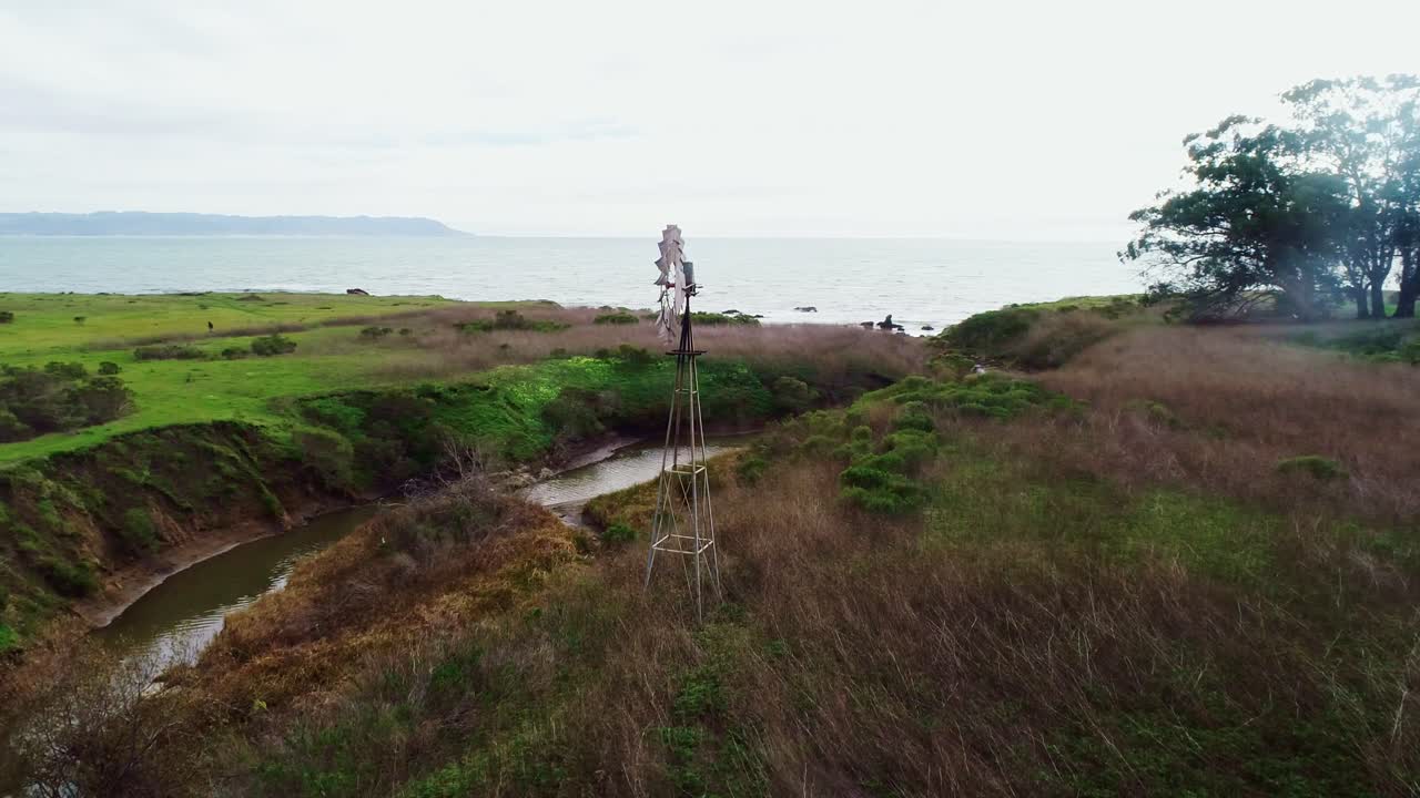 Windmill turning beside a winding coastal creek and green fields in California