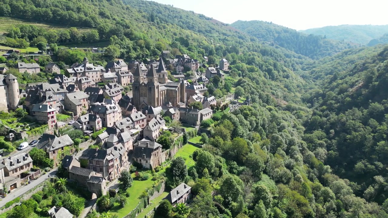 Veduta aerea con drone nella campagna francese di una piccola vecchia città medievale e una cattedrale circondate da una verde foresta montana, con panoramica a Conques