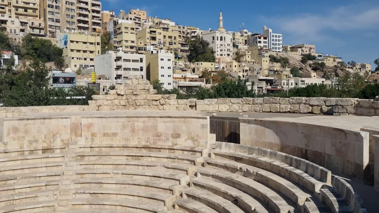 All'interno del teatro romano Odeon di Amman, le antiche gradinate in pietra e l'architettura classica ben conservata contrastano con un tipico quartiere collinare sotto un cielo azzurro limpido e una luce diurna brillante.