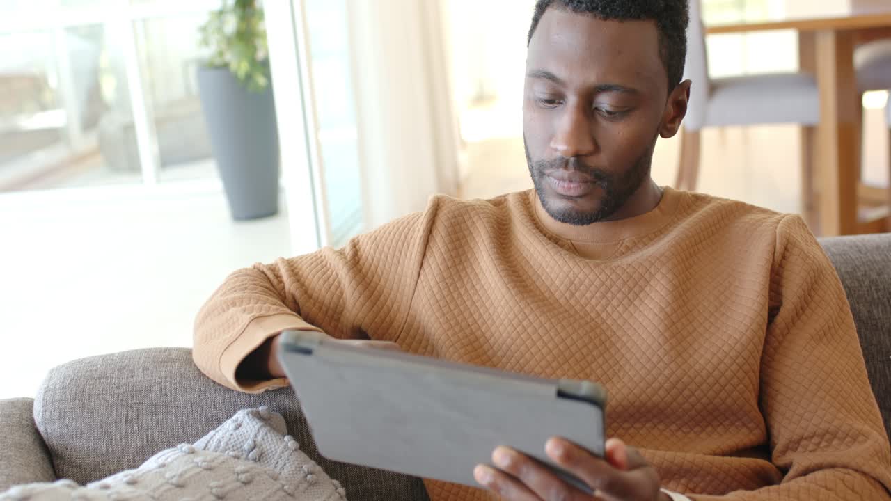 Lifting tablet, African American man tapping touchscreen and browsing content on couch by window
