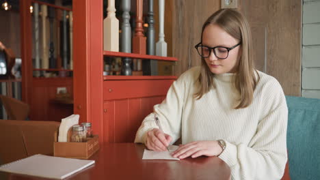 woman in glasses and white sweater seated in cozy cafe reaching toward notebook on wooden table with condiment holder and pen nearby, warm ambiance and striped background