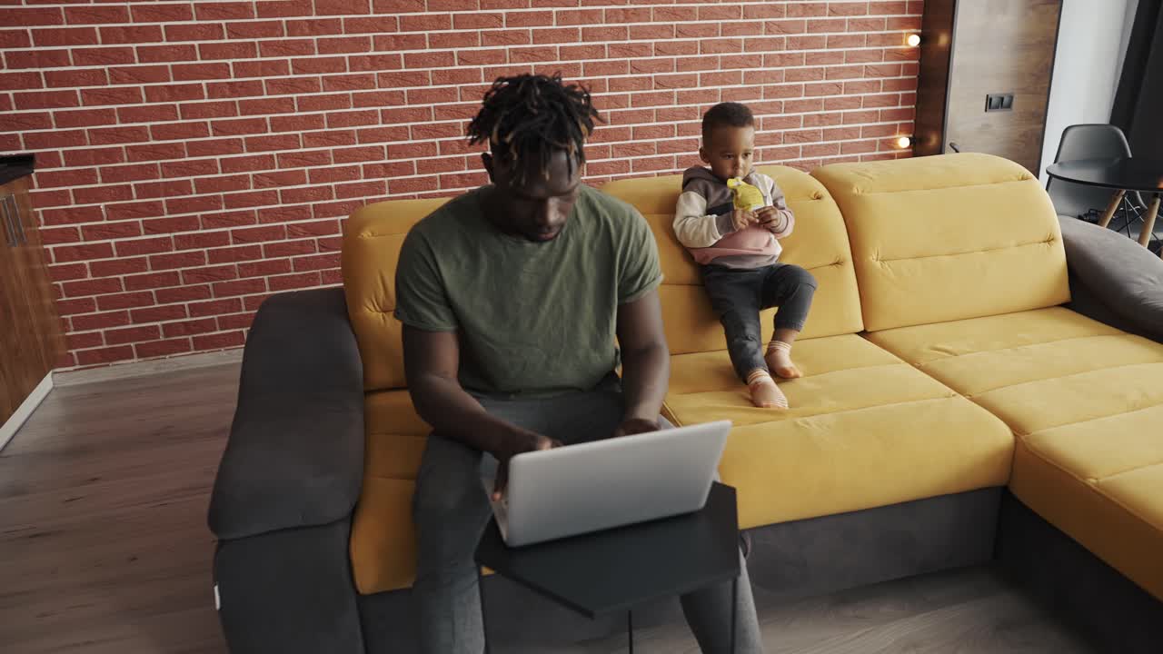 Busy young african father doing freelance work on laptop and babysitting young child at home