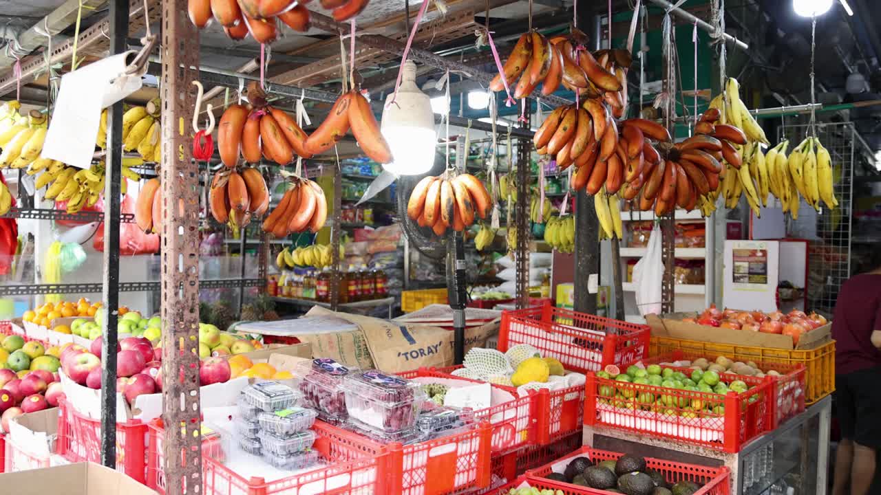 Red bananas and yellow bananas hang above crates of assorted fruits in a brightly lit, bustling Singapore market. Static camera, vibrant colors, indoor setting