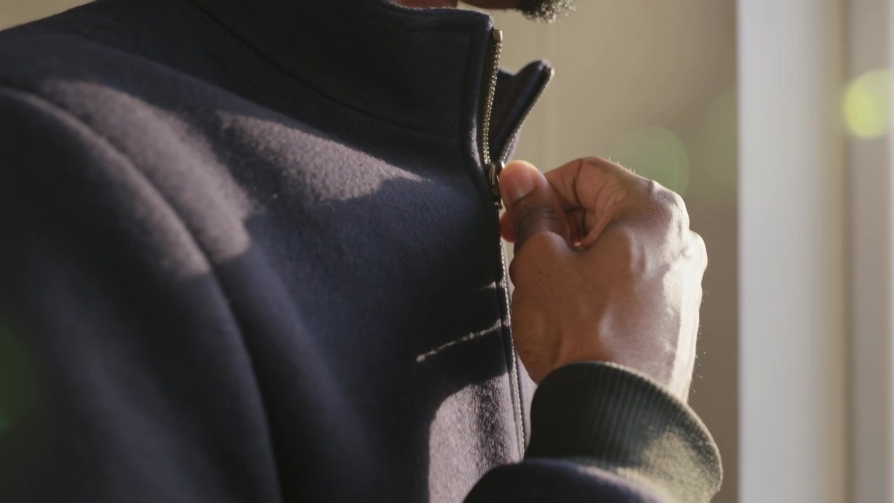 Reaching adult man gripping metal zipper pull at window, pulling dark jacket collar up to fasten