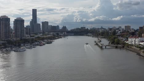 Toma-De-Drones-En-ángulo-Alto-Con-Un-Barco-Gato-De-La-Ciudad-En-El-Río-Brisbane-En-New-Farm,-Siguiendo-A-Un-Grupo-De-Ciclistas-Mientras-Recorren-El-Paseo-Del-Río-New-Farm