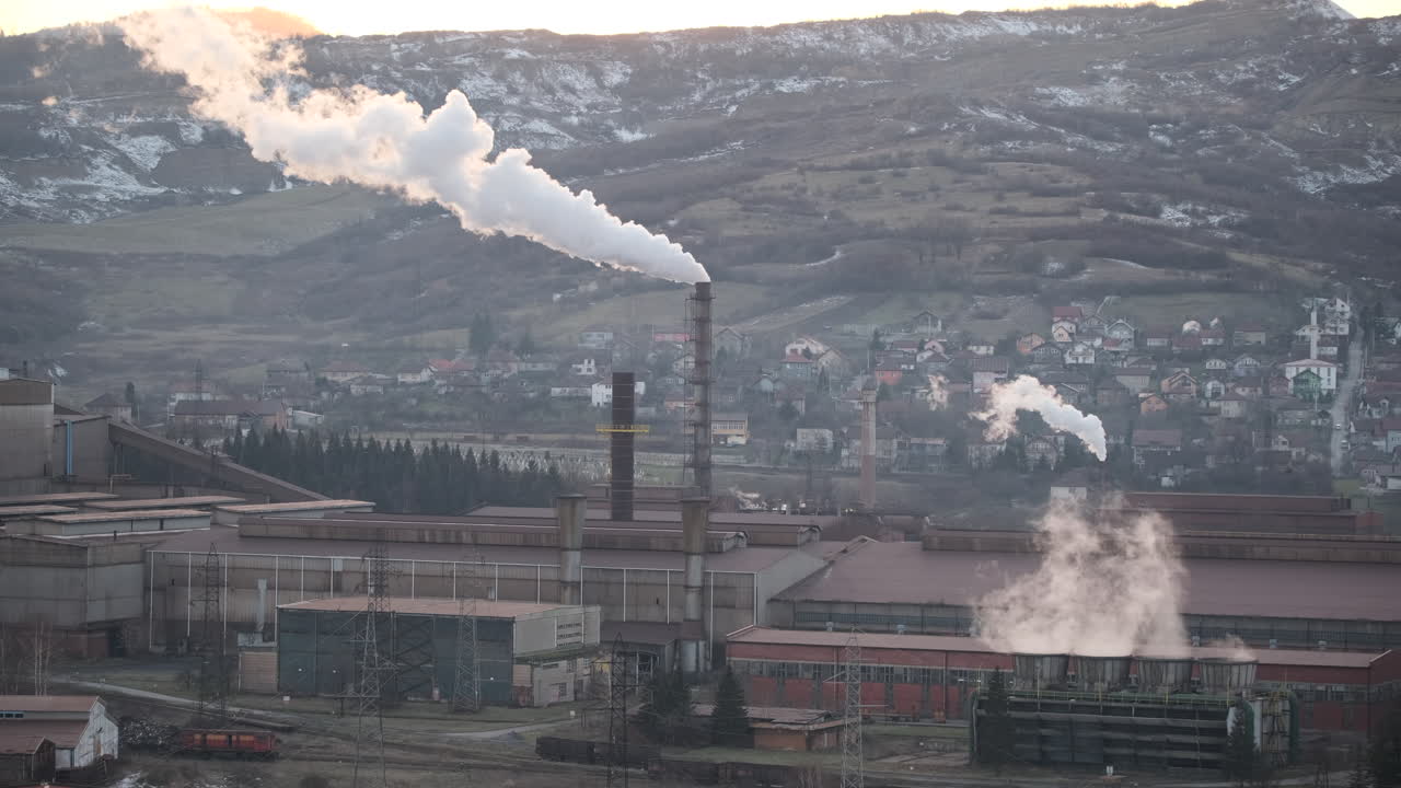 una columna de vapor se eleva prominentemente desde una chimenea alta en una planta industrial en un valle, con áreas residenciales y colinas cubertas de nieve en el fondo