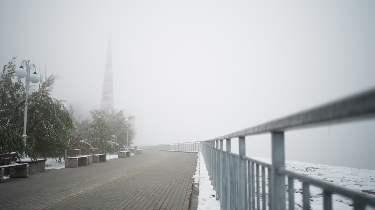 안개가 가득한 공원 벤치, 얼어붙은 나무, 철의 레일링, 조용한 산책로를 따라 빛의 기둥을 는 첫 눈이 내리는 겨울 도시 풍경.