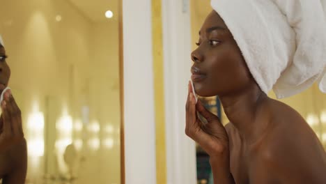 African-american-woman-with-towel-using-pad-on-her-face-and-watching-in-mirror-in-bathroom